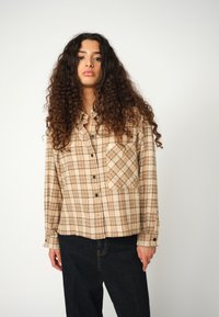 Giovane donna con lunghi capelli ricci, che indossa una camicia a quadri beige con bottoni e jeans in denim scuro, di fronte a uno sfondo bianco semplice.