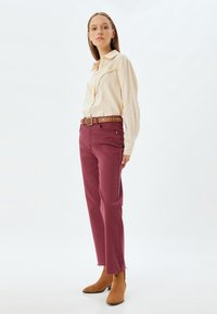 Blusa di crema chiara con dettaglio di volant, abbinata a pantaloni dritti bordeaux e una cintura marrone con borchie. Stivaletti marroni completano il look.