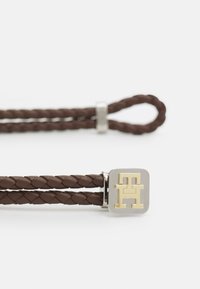 Il braccialetto in pelle intrecciata marrone presenta una chiusura in argento con un dettaglio del logo dorato. La texture liscia contrasta con il design intrecciato.