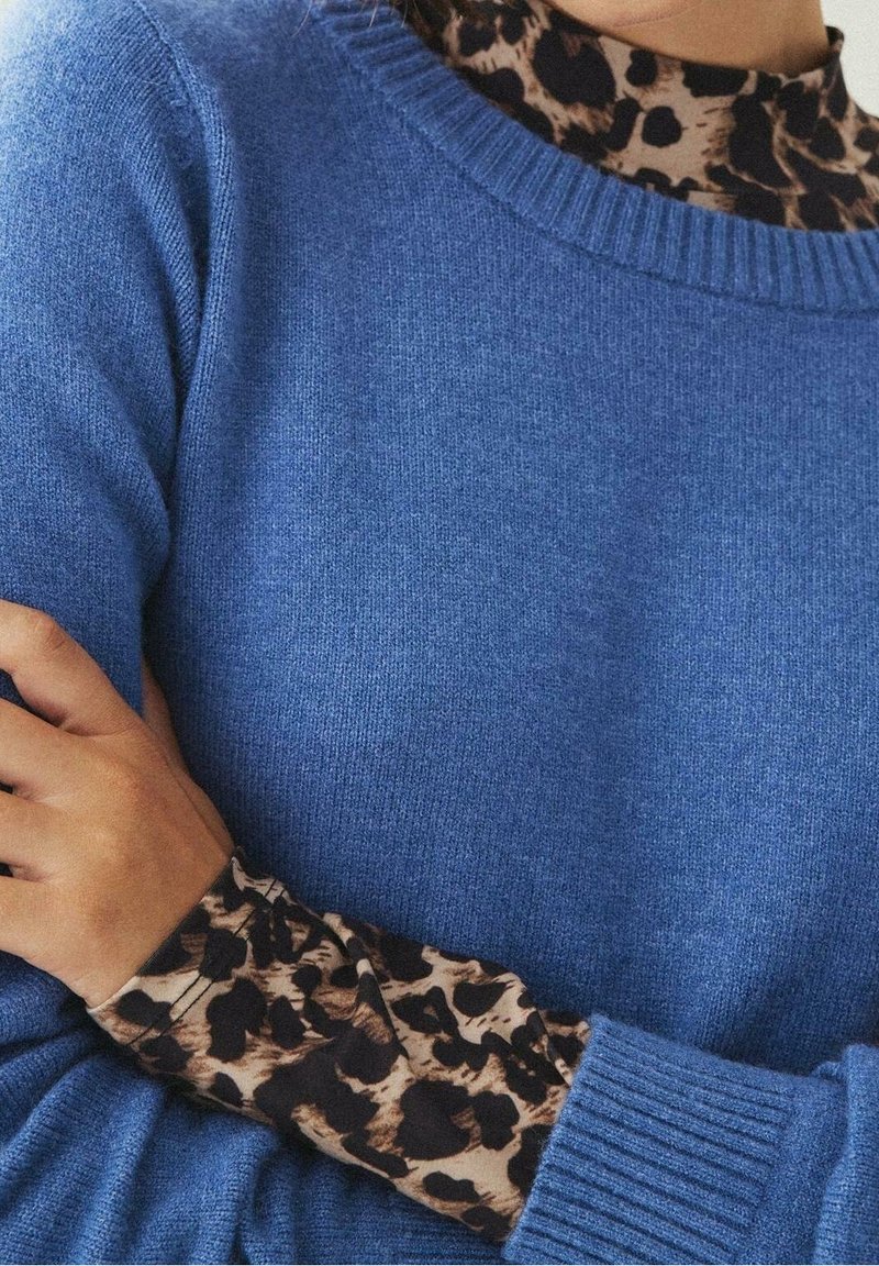 Personne portant un pull bleu en tricot sur une chemise à manches longues à motif léopard, les bras croisés sur la poitrine.