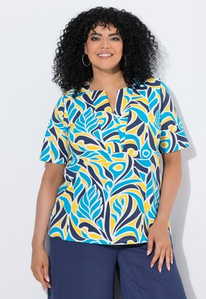 Vrouw met krullend haar die een kleurrijke kortemouwige top met patroon en donkerblauwe broek draagt, glimlachend tegen een effen achtergrond.