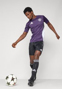 Lilla og hvid fodboldtrøje med korte ærmer, sorte shorts og sorte fodboldstøvler. Spiller dribler en mønstret fodbold.