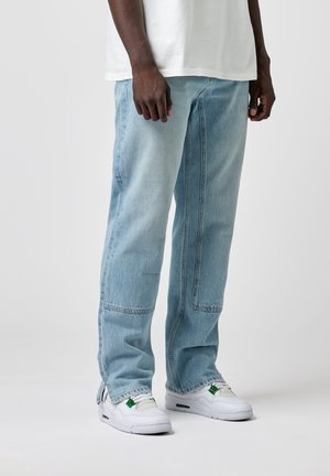 Homme portant un jean patchwork bleu clair et des baskets blanches avec des accents verts, debout devant un fond uni.