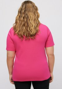 Korte mouwen roze t-shirt met een zachte textuur, afgeronde onderrand en een ontspannen pasvorm, gecombineerd met zwarte onderkleding op de achtergrond.