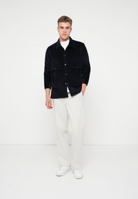 Chaqueta de pana azul marino con dos bolsillos frontales, cierre de botón y cuello con solapa. Llevada sobre una camisa blanca con pantalones crema y zapatillas blancas.