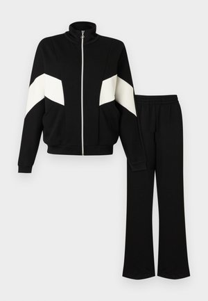 Survêtement noir avec des bandes en chevron blanches sur les manches, veste zippée à col montant, et pantalon assorti à taille élastique.
