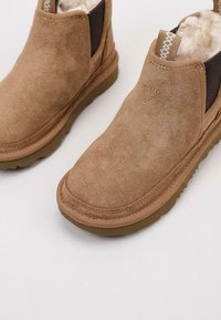 Botas de tobillo de ante color arena con un interior suave parecido al de la piel de un animal, paneles laterales elásticos y una suela de goma marrón; presenta detalles de costura en la parte superior.