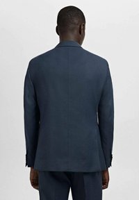 Mężczyzna z krótkimi włosami ubrany w granatowy żakiet i pasujące spodnie, stojący tyłem na jednolitym jasnym tle.