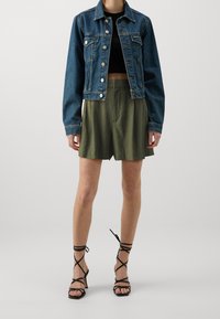 Nainen, joka on pukeutunut tummansinisestä denim-jakusta, mustasta crop-topista, oliivinvihreistä shortseista ja mustista nilkkakorkokengistä, seisoo yksinkertaisella taustalla.