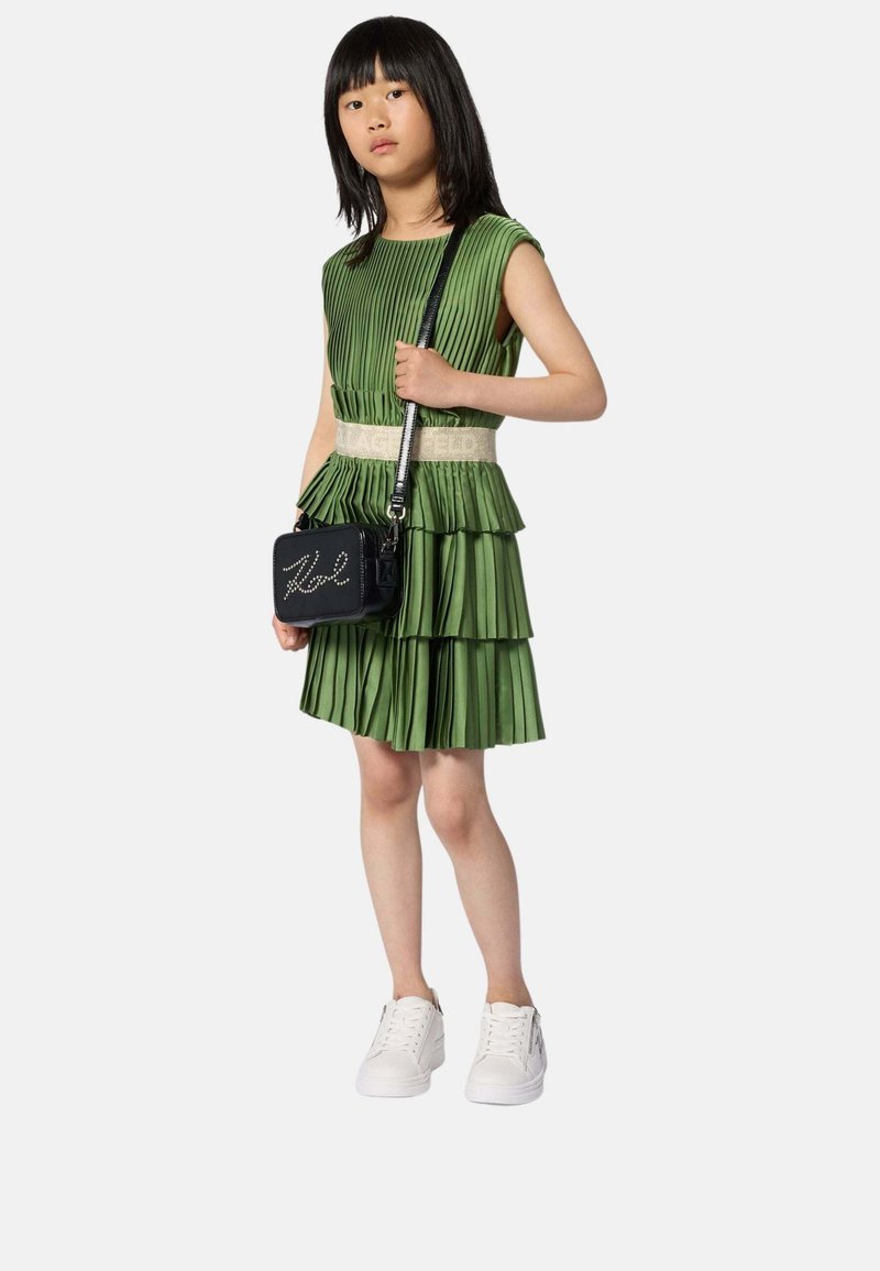 Jeune fille portant une robe verte plissée à volants avec une ceinture beige, des baskets blanches, et tenant un sac bandoulière noir avec des lettres cloutées.