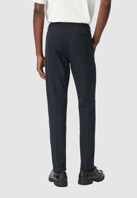 Marineblauwe broek met een getailleerde pasvorm, elastische tailleband en één achterzak. Het materiaal is glad met een subtiele glans, gecombineerd met zwarte schoenen.