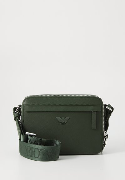 Sac à bandoulière en simili cuir vert avec fermeture zippée, poche avant, logo embossé et bandoulière en tissu réglable. Texture lisse et forme rectangulaire.