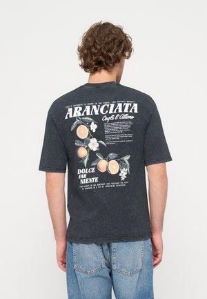 Mand iført en sort t-shirt med grafik af en appelsin og en hvid blomst samt italiensk tekst på ryggen, kombineret med blå jeans.