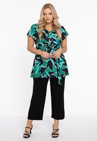 Bluse mit Blumenmuster in Grün, Blau und Schwarz, mit kurzen Rüscheärmeln und einem Bindegürtel. Kombiniert mit schwarzen Culotte-Hosen und Riemchensandalen.