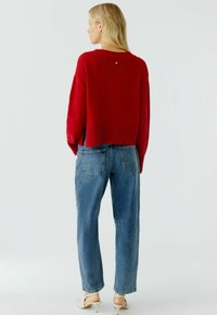 Rode gebreide trui met geribbelde zoom, getoond met lichtblauwe hooggelegen jeans. Opvallend detail: kleine accent aan de achterkant van de halslijn.