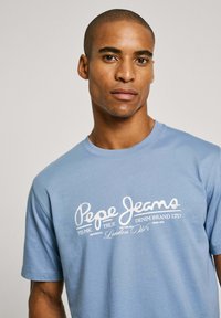 Svijetloplava pamučna majica s bijelim printanim logotipom koji glasi "Pepe Jeans" s dodatnim tekstom ispod. Kratki rukavi, okrugli izrez.