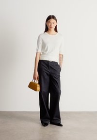 Bela majica s kratkimi rokavi, široke temno modre kavbojke, sijoči črni čevlji in majhna rumena torbica, modelka stoji pred svetlim ozadjem.