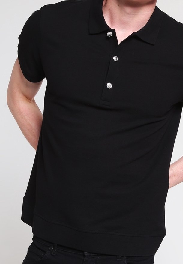 Homme portant un polo noir uni à manches courtes avec trois boutons, debout les mains dans le dos, sur un fond clair.