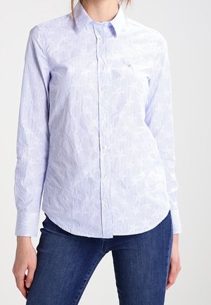 Kvinde iført lyseblå og hvide vertikalt striped skjorte med kraver, kombineret med mørkeblå jeans, mod en ensfarvet baggrund.