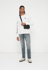 Hvit skjorte med lange ermer og svart logo, grå denimjeans og beige sneakers. Svart crossbody-veske med teksturert design.