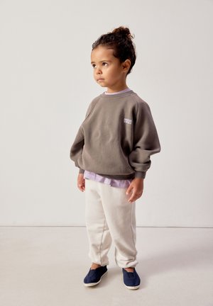 Jaunas vaikas su garbanotais plaukais surištais į kuoduką, dėvi pilką džemperį, po juo šviesiai violetinę palaidinę, baltas kelnes ir tamsiai mėlynas slip-on tipo batus, stovi patalpose.