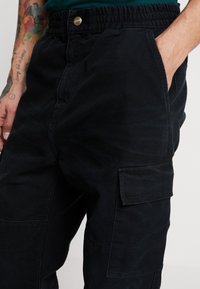 Donkerblauwe cargobroek van stevig materiaal, met een elastische tailleband, zijzakken en een patchzak op de dij.