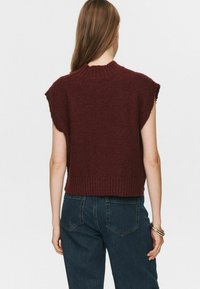 Moteris su ilgais plaukais, vilkinti be rankovių tamsiai raudoną megztą liemenę ir tamsiai mėlynas džinsines kelnes, atsisukusi nugarą, su apyrankėmis dešinėje riešo pusėje.