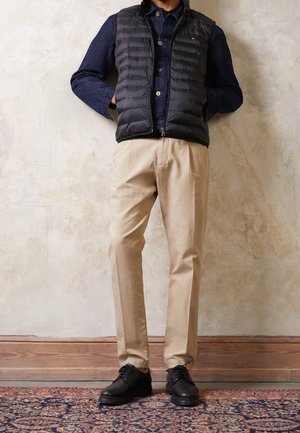 Mann trägt schwarze Steppweste, dunkelblaues Hemd mit Knöpfen, beige Hose und schwarze Schuhe, steht vor einer strukturierten Wand auf einem gemusterten Teppich.