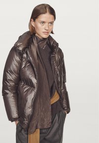 Brązowa kurtka puff z błyszczącym wykończeniem, wysokim kołnierzem i zamkiem błyskawicznym z przodu. Połączona z ciemnym swetrem i prążkowanym szalikiem, posiadającym brązowy pasek.