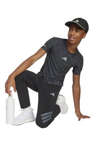 T-shirt de sport noir à motifs et pantalon noir avec trois bandes grises. Modèle tenant une bouteille d'eau blanche, portant des chaussures blanches et une casquette noire.