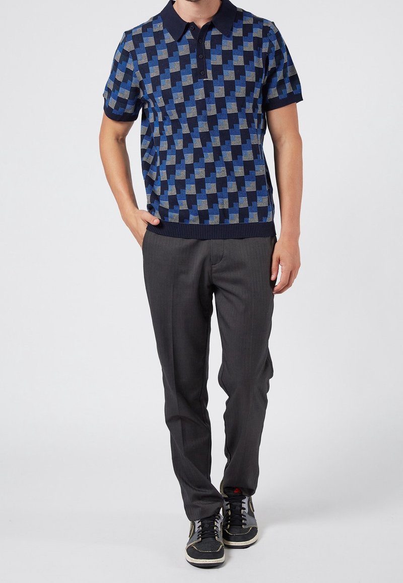 Homme portant un polo manches courtes à carreaux bleu et noir, pantalon gris foncé et baskets noires, debout avec une main dans la poche.