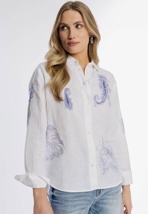 Kobieta o długich blond włosach, ubrana w białą, długorękawową koszulę zapinaną na guziki, ozdobioną niebieskimi motywami kwiatowymi i paisley, w połączeniu z jasnoniebieskimi dżinsami.