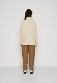 Top din fleece bej cu guler înalt, asortat cu pantaloni cargo maro cu buzunare laterale și adidași argintii cu talpă albă.