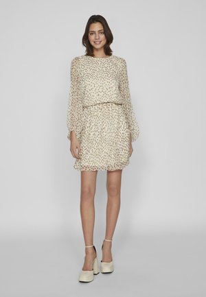 Beige jurk met een losse pasvorm, voorzien van een stippenpatroon, lange mouwen en een verzamelde taille. Gepaard met witte platform schoenen.