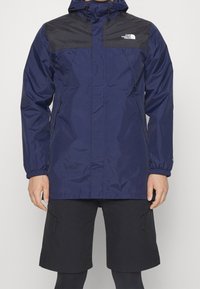 Navy wodoodporna kurtka z czarnymi akcentami na ramionach, regulowanym kapturem, elastycznymi mankietami i centralnym zamkiem. Połączona z czarnymi szortami cargo.