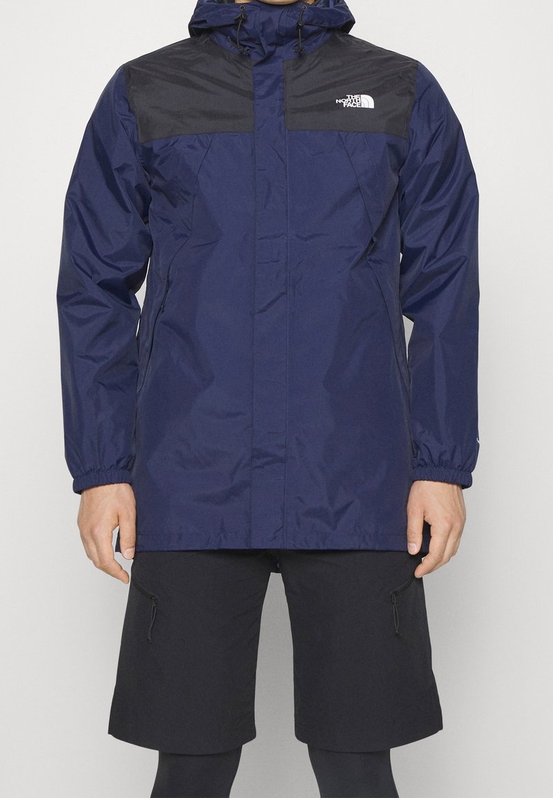 Navy wodoodporna kurtka z czarnymi akcentami na ramionach, regulowanym kapturem, elastycznymi mankietami i centralnym zamkiem. Połączona z czarnymi szortami cargo.