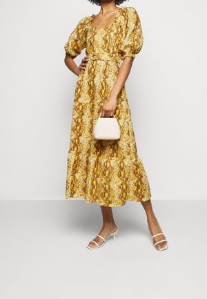 Kobieta w żółto-brązowej, midi sukience w wzory z bufkami na rękawach, trzymająca małą beżową torebkę i nosząca białe sandały na obcasie.