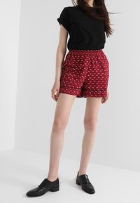 Röda shorts med vitt hjärtmönster, elastisk midja och uppvikta fållar, kombinerade med en svart kortärmad topp och svarta skor.