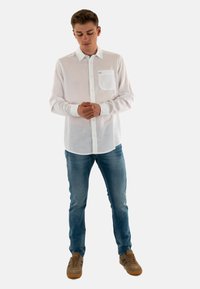 Chemise blanche à manches longues, avec un poche, texture légère, associée à un jean bleu et des baskets marron. Fond neutre.
