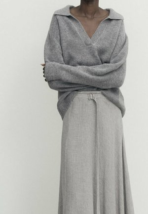 Personne portant un pull ample gris à col en V avec manches longues, croisé sur une jupe taille haute gris clair texturée avec une petite boucle de ceinture.