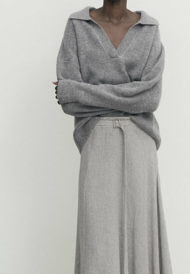 Personne portant un pull ample gris à col en V avec manches longues, croisé sur une jupe taille haute gris clair texturée avec une petite boucle de ceinture.