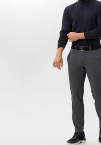 Man wearing black turtleneck, gray checkered trousers, black belt, and black sneakers, adjusting his left sleeve against a plain background.