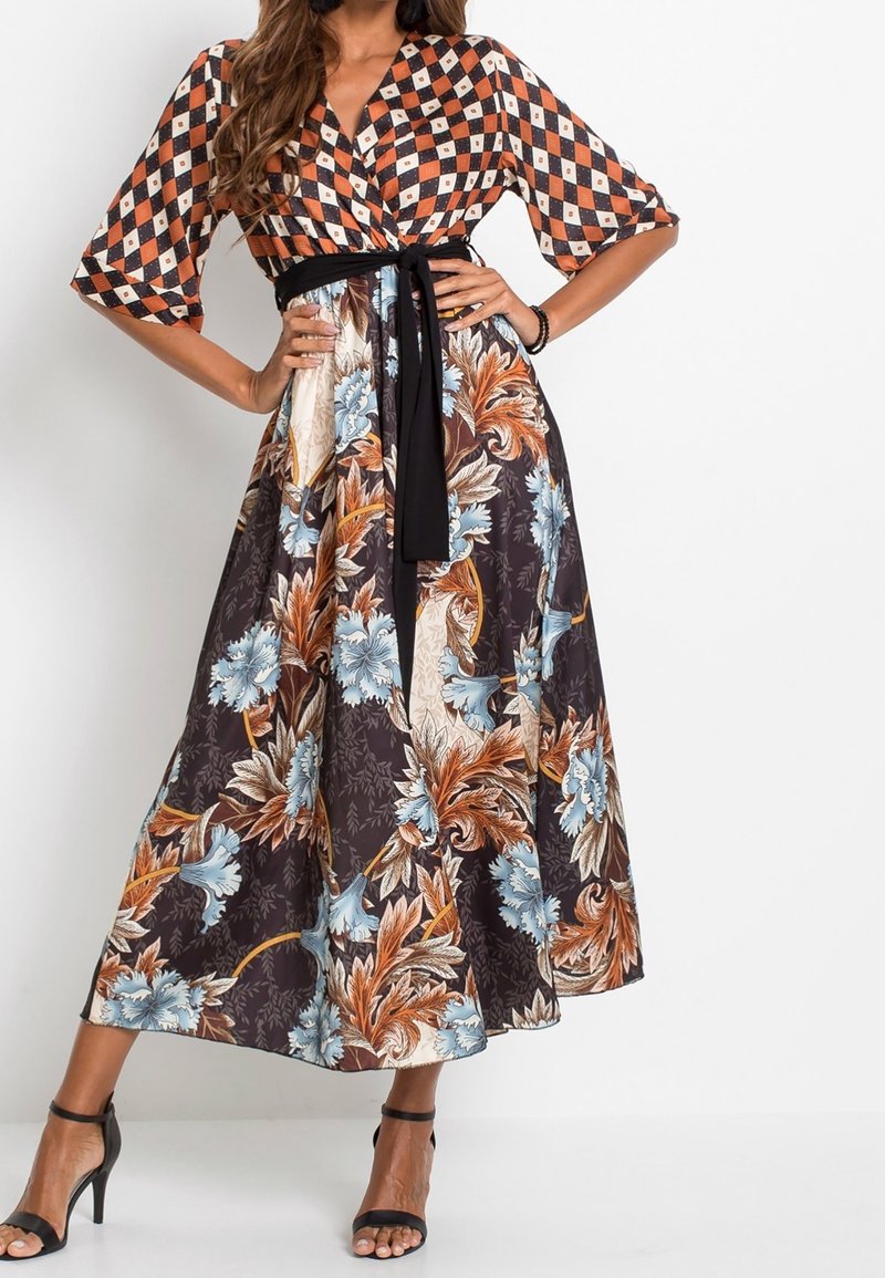 Donna che indossa un vestito a portafoglio con parte superiore a scacchi neri, bianchi e arancioni e una gonna a motivo floreale nelle tonalità blu, marrone e beige, con tacchi neri.
