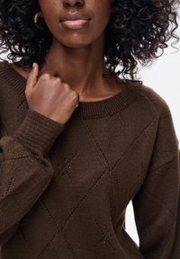 Kobieta o kręconych włosach ubrana w brązowy sweter dziergany z diamentowymi wzorami, trzymająca sweter za kołnierz jedną ręką.