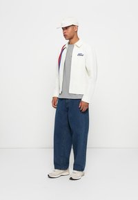 Man wearing white Ford jacket with red and blue stripe, gray shirt, loose blue jeans, white sneakers, and white cap standing against plain white background.