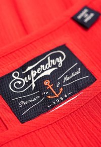 Röd tygbakgrund med en svart vävd etikett med märket "Superdry", vit text i skrivstil och en orange ankarsymbol.