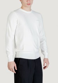 Pull en maille blanc à manches longues avec poignets et ourlet côtelés, arborant un petit logo discret sur la poitrine et un col rond classique.