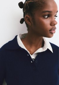 Polo shirt in maglia di colore blu navy con colletto, sovrapposto a una camicia a righe bianca e grigia. Presenta tre bottoni e una texture morbida.