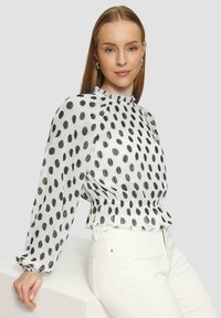 Witte blouse met zwarte stippen. Beschikt over een hoge halslijn, lange mouwen met elastische manchetten en een gerimpelde onderkant. Draagt met witte broek.