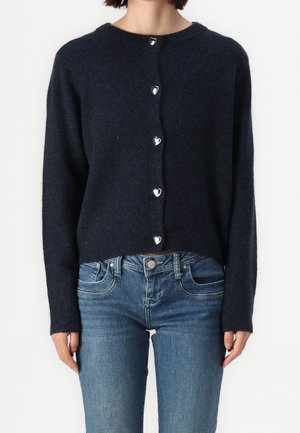 Femme portant un cardigan duveteux bleu marine avec des boutons en forme de cœur, assorti à un jean de lavage moyen, sur un fond uni.